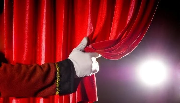 Een hand opent een rood theatergordijn in het licht van een podiumspot.